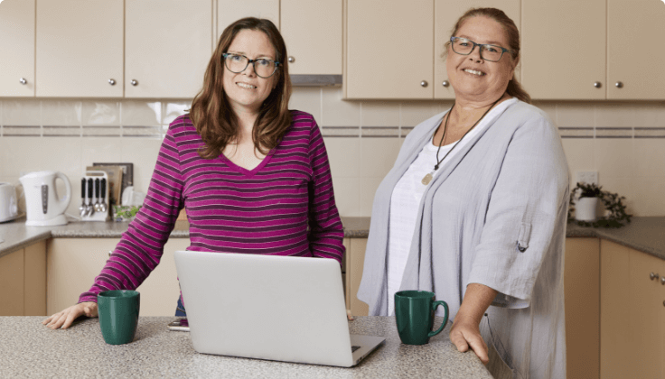 Two women (1 presumed to have an intellectual or cognitive disability and the other being a support worker) which are standing in the kitchen with computer and coffee mugs on the kitchen bench.