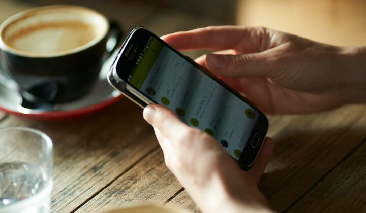 In focus image of hands holding a mobile phone with cup of coffee and cup of water placed on the table.