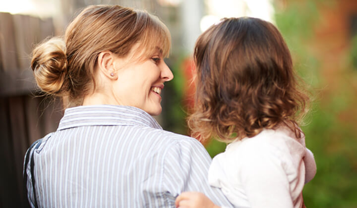 Light skin woman smiling and holding young daughter up close to her head and shoulder. Photo taken from behind and young daughters face cannot be seen.