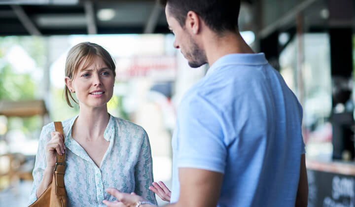 Light skin woman with concerned look on her face standing in close proximity to her partner who is the perpetrator of technology facilitated abuse against her.